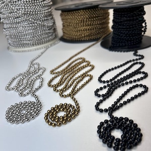 May include: Three spools of ball chain in silver, brass, and black. Strands of each color are laid out, showcasing the bead-like links. The chains are likely used for jewelry, crafts, or decorative purposes.
