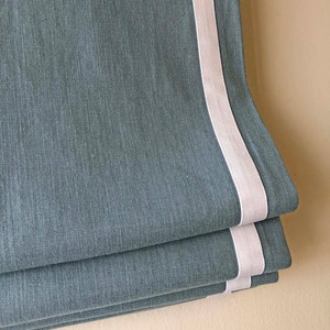 May include: A blue linen roman shade with a white trim. The shade is folded up, showing the layers of fabric.