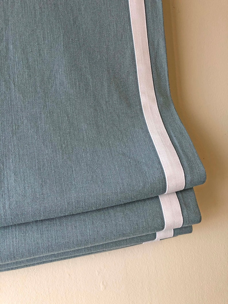 May include: A blue linen roman shade with a white trim. The shade is folded up, showing the layers of fabric.