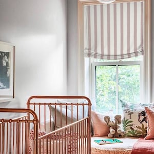 K&ouml;nnte beinhalten: Zwei Kupfer-Babybetten mit wei&szlig;en Bettw&auml;sche und Kissen. Die Babybetten stehen neben einem Fenster mit einem rosa-wei&szlig;en gestreiften r&ouml;mischen Rollo. Das Fenster blickt auf einen gr&uuml;nen Baum.