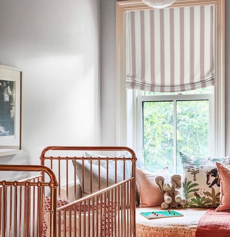 K&ouml;nnte beinhalten: Zwei Kupfer-Babybetten mit wei&szlig;en Bettw&auml;sche und Kissen. Die Babybetten stehen neben einem Fenster mit einem rosa-wei&szlig;en gestreiften r&ouml;mischen Rollo. Das Fenster blickt auf einen gr&uuml;nen Baum.