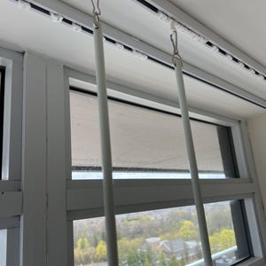May include: Two white curtain wands hang vertically from a white ceiling-mounted track system. Each wand is attached to the track by a small metal hook and a white plastic slider. The track features a series of evenly spaced white plastic gliders.