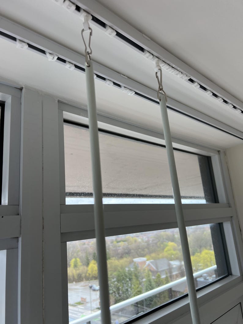 May include: Two white curtain wands hang vertically from a white ceiling-mounted track system. Each wand is attached to the track by a small metal hook and a white plastic slider. The track features a series of evenly spaced white plastic gliders.