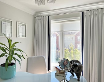 May include: A white office with a large window, white curtains with navy blue trim, a potted plant, a white chair, a globe, and a gray cat.