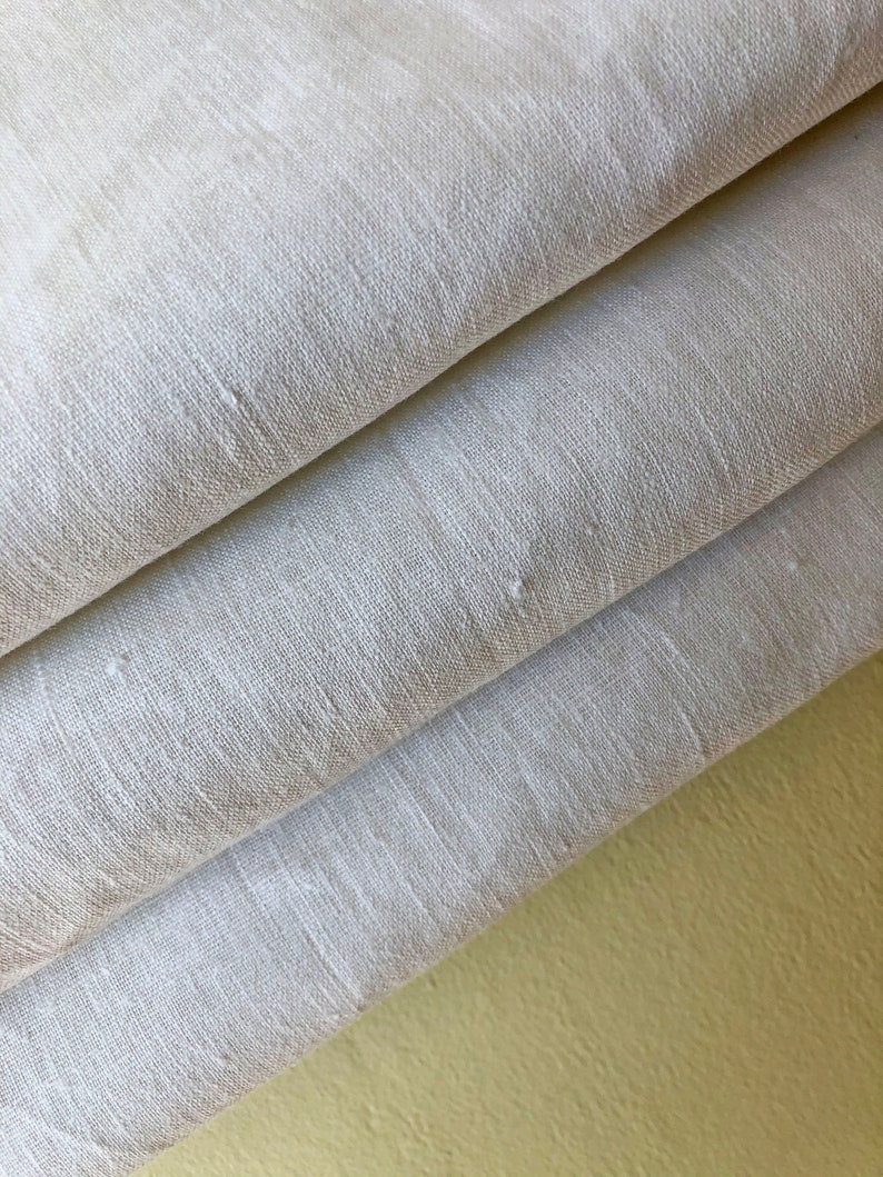 May include: A stack of three white linen fabric panels. The fabric is smooth and has a subtle texture. The panels are folded in thirds and the edges are visible. The fabric is a light cream color.