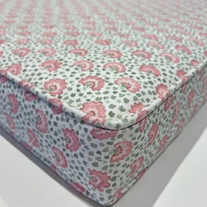 May include: A white cushion with a pink and green floral pattern. The cushion has a white border and is made of a soft fabric.