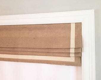 May include: A brown roman shade with a white trim. The shade is mounted in a white window frame.