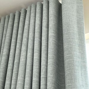 May include: A floor-to-ceiling curtain panel in a light gray color with a visible woven fabric texture. The curtain is hung from a white track system at the top, creating deep, uniform vertical folds that extend down the length of the fabric.