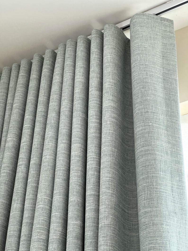 May include: A floor-to-ceiling curtain panel in a light gray color with a visible woven fabric texture. The curtain is hung from a white track system at the top, creating deep, uniform vertical folds that extend down the length of the fabric.