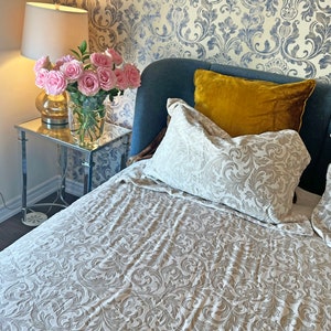 May include: A bed with a white and gray patterned duvet cover and two pillows. The headboard is a dark blue velvet. A yellow velvet pillow is on top of a white pillow. A side table with a lamp and a vase of pink roses is in the background.