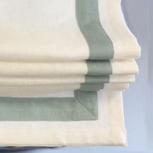 May include: White roman shade with a green trim. The shade is folded up, showing the layers of fabric.