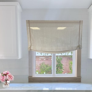 May include: A white kitchen with white cabinets and a window with a linen Roman blind. The window is above a white worktop with a vase of pink flowers.