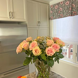 May include: A bouquet of pink roses in a clear glass vase on a granite countertop in a kitchen. The roses are arranged in a vase with green stems and leaves. The kitchen has white cabinets and a stainless steel refrigerator.