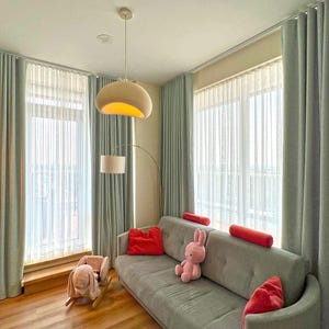 May include: A gray sofa with red throw pillows and a pink rabbit plush toy, positioned in a room with floor-to-ceiling light gray curtains, a white pendant light, a floor lamp, and a wooden rocking toy shaped like a rabbit on the floor.