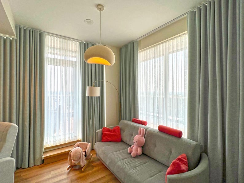 May include: A gray sofa with red throw pillows and a pink rabbit plush toy, positioned in a room with floor-to-ceiling light gray curtains, a white pendant light, a floor lamp, and a wooden rocking toy shaped like a rabbit on the floor.