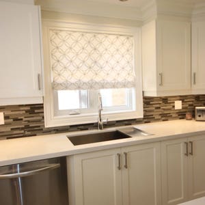 May include: White kitchen cabinets with a stainless steel dishwasher and a white countertop with a sink. The window has a white roman shade with a geometric pattern.