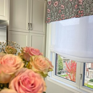 May include: A white window with a floral valance and a white roman shade. The window is looking out onto a street with trees and buildings. A bouquet of pink roses is in the foreground.