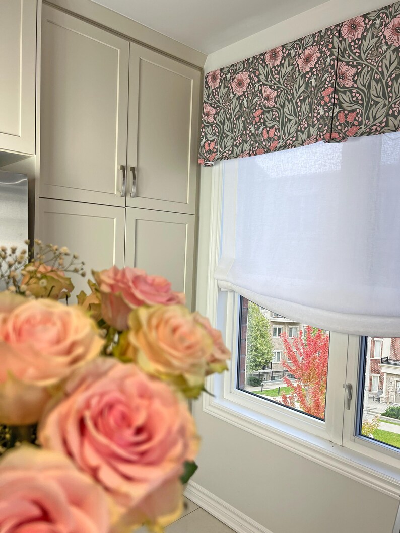 May include: A white window with a floral valance and a white roman shade. The window is looking out onto a street with trees and buildings. A bouquet of pink roses is in the foreground.