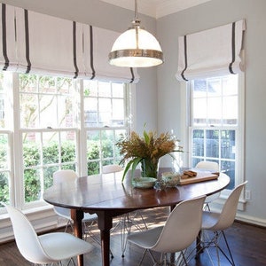 May include: A dining room with a wooden table and white chairs. The windows have white roman shades with black stripes. There are plants on the table.