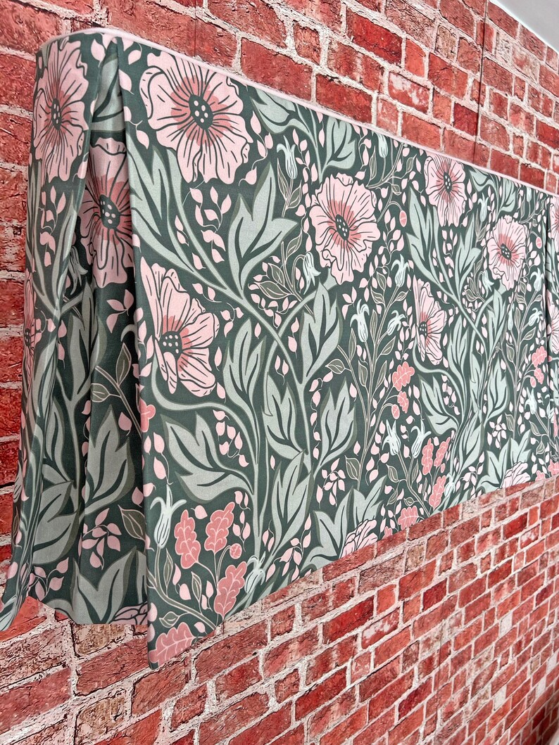 May include: A green and pink floral patterned fabric valance with a pleated edge. The valance is hanging on a brick wall.