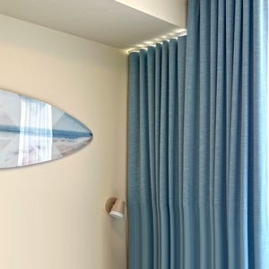 May include: Floor-to-ceiling light blue curtains with a vertical pleated texture hang from a track. A surfboard-shaped wall art piece featuring a beach scene with white and blue tones is mounted on the adjacent cream-colored wall.