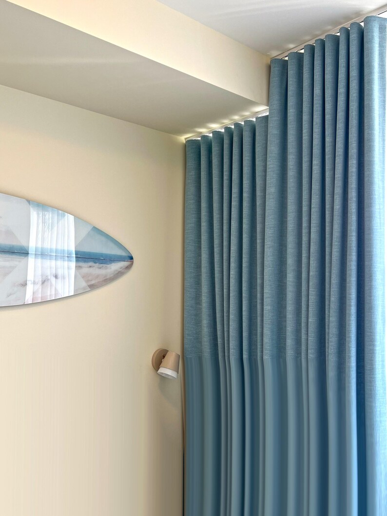 May include: Floor-to-ceiling light blue curtains with a vertical pleated texture hang from a track. A surfboard-shaped wall art piece featuring a beach scene with white and blue tones is mounted on the adjacent cream-colored wall.