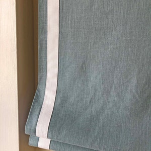 May include: A gray roman shade with a white trim. The shade is made of a textured fabric.