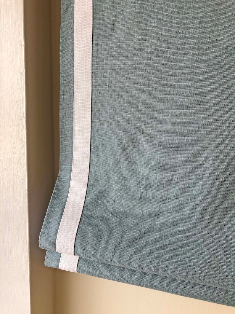 May include: A gray roman shade with a white trim. The shade is made of a textured fabric.