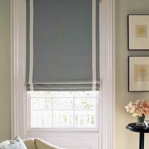 May include: A white window frame with a gray roman shade with white trim. The shade is pulled up to reveal a window with white panes.