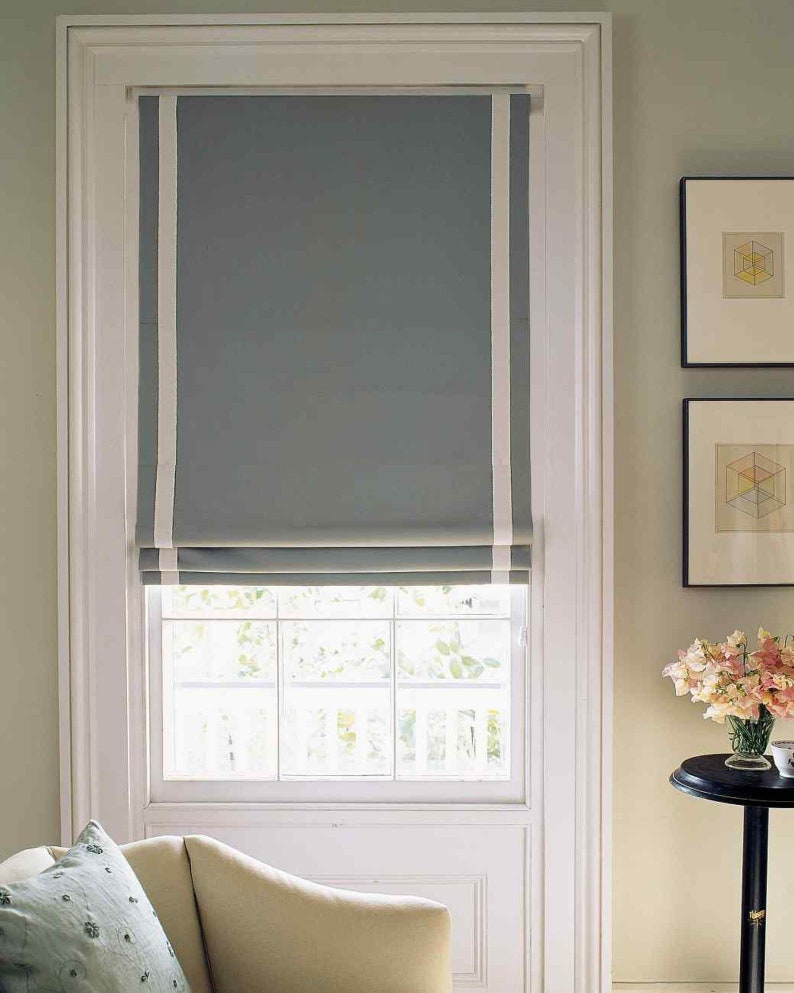 May include: A white window frame with a gray roman shade with white trim. The shade is pulled up to reveal a window with white panes.