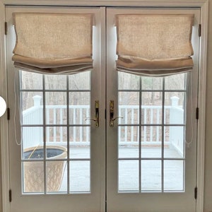 May include: Two white french doors with gold door handles. The doors have a tan roman shade on each window. The doors open to a view of a white wooden deck with a potted plant.