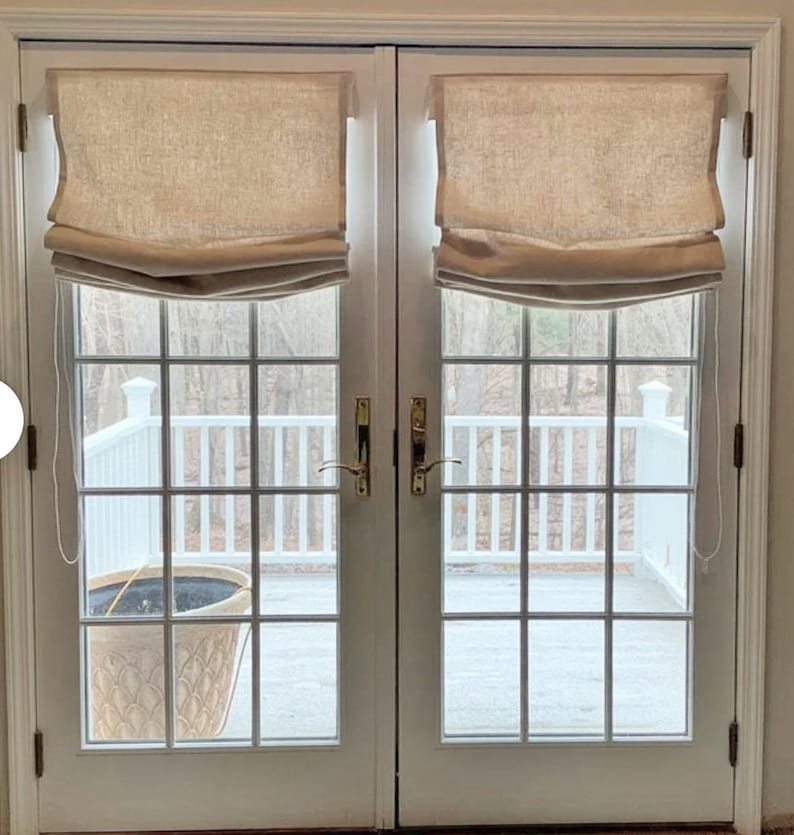 May include: Two white french doors with gold door handles. The doors have a tan roman shade on each window. The doors open to a view of a white wooden deck with a potted plant.
