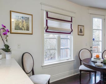 May include: A white kitchen with a window featuring a white roman shade with a burgundy trim. Two chairs are positioned in front of a table with a silver pitcher and a plate of fruit.