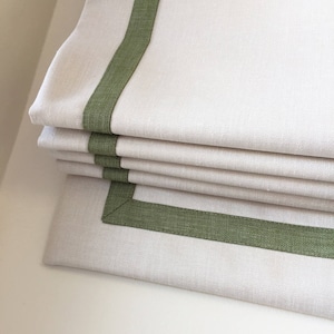 May include: A white roman shade with a green trim. The shade is folded up, showing the layers of fabric.