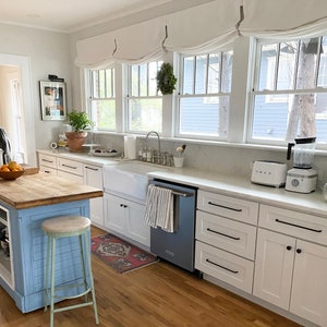 May include: A white kitchen with a farmhouse sink, white cabinets, and a blue dishwasher. The kitchen has a wood countertop and a wooden floor. There is a wreath hanging on the window. There is a toaster oven and a blender on the counter.