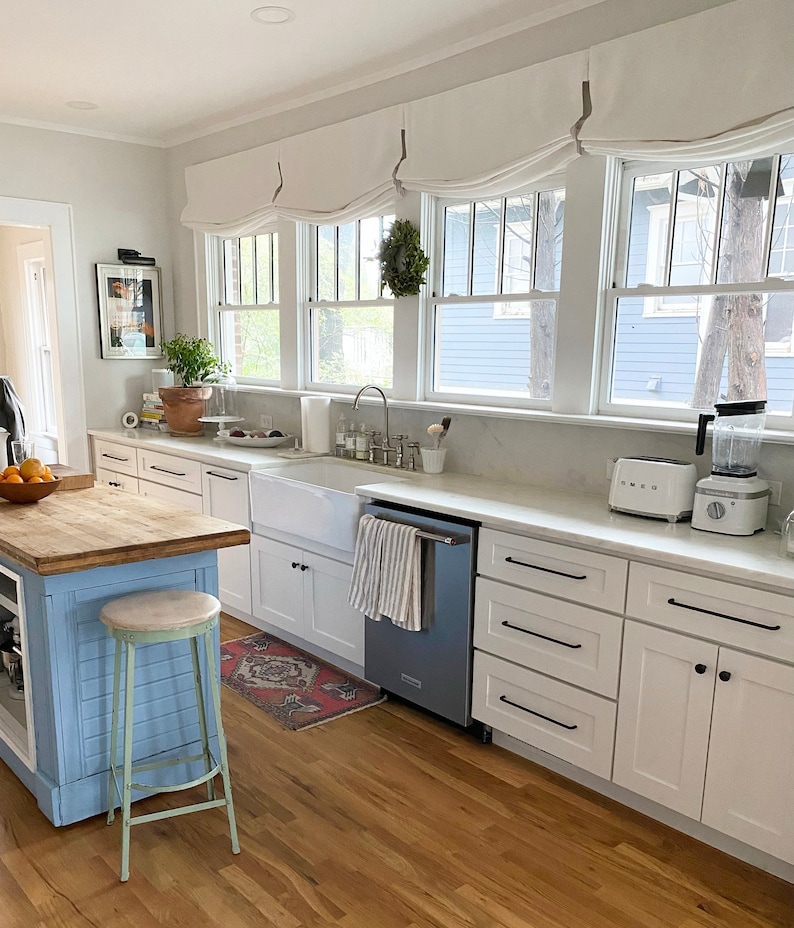 May include: A white kitchen with a farmhouse sink, white cabinets, and a blue dishwasher. The kitchen has a wood countertop and a wooden floor. There is a wreath hanging on the window. There is a toaster oven and a blender on the counter.