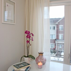 May include: A white table with a pink vase, a lit candle, and a stack of books. A pink orchid plant is in a white pot on the table. The table is in front of a window with white curtains and a view of a building.
