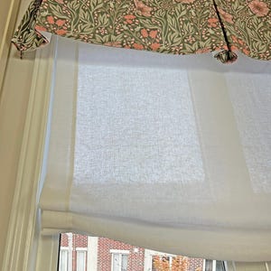May include: White roman shade with a floral valance in shades of pink and green. The shade is pulled up to reveal a window with a view of a brick building and trees.