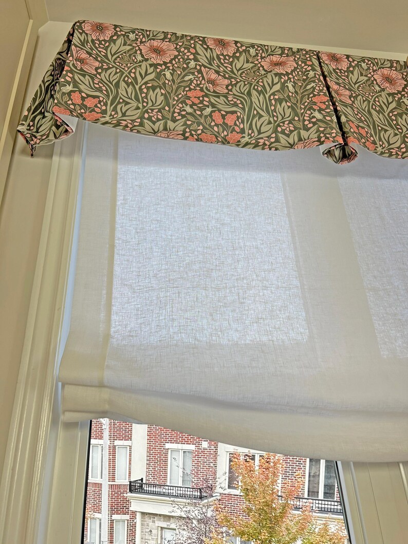May include: White roman shade with a floral valance in shades of pink and green. The shade is pulled up to reveal a window with a view of a brick building and trees.
