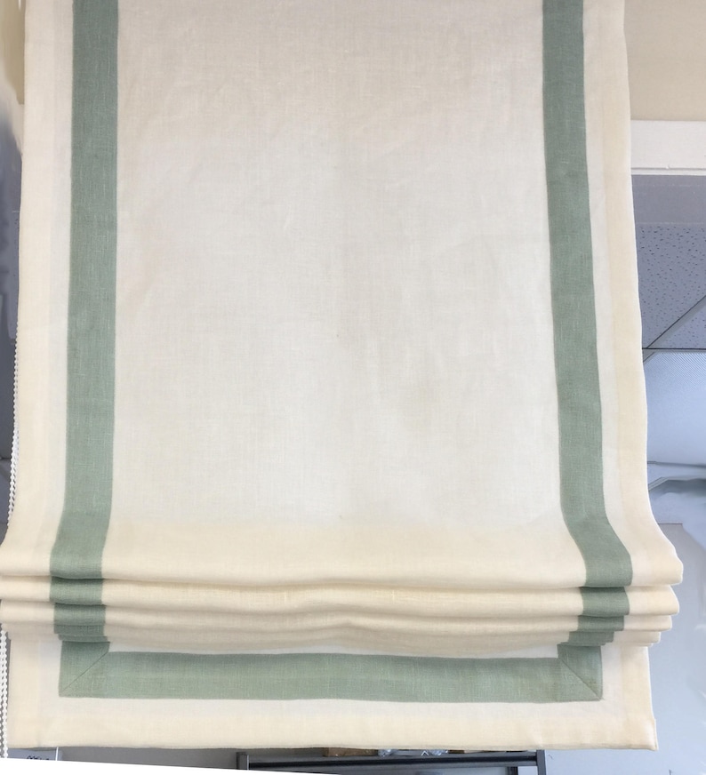 May include: A white roman shade with a green border. The shade is pulled up and folded, showing the layers of fabric.
