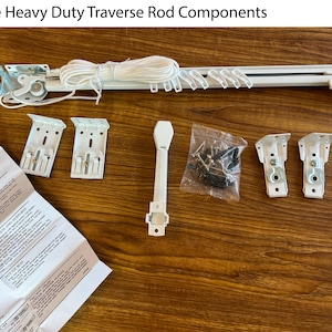 May include: A collection of single heavy-duty traverse rod components. The set includes a white traverse rod, mounting brackets, a cord, screws, and an instruction sheet. The components are arranged on a brown wooden surface.