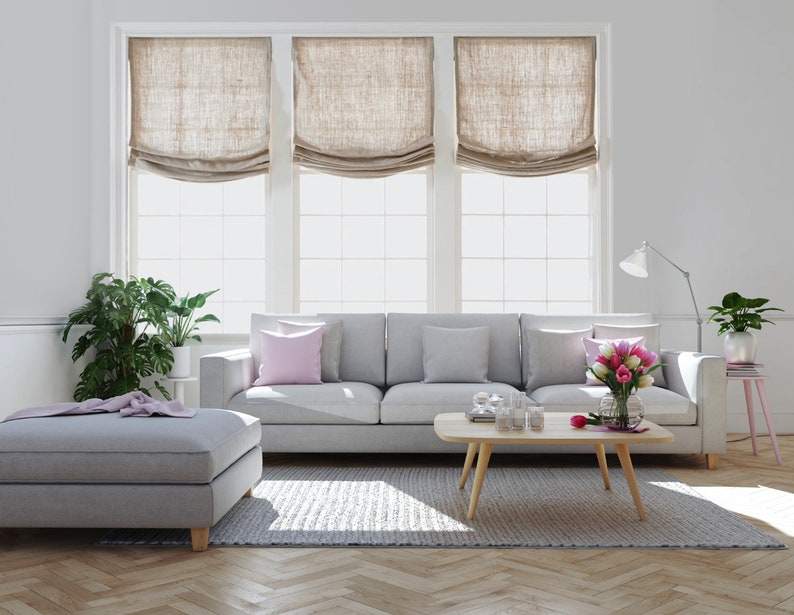 May include: A gray sofa with three pink throw pillows, a gray ottoman, a wooden coffee table with clear glass candle holders, a vase of pink flowers, a green plant in a white pot on a pink side table, and a floor lamp with a white shade. The room has white walls and a wood floor with a herringbone pattern.