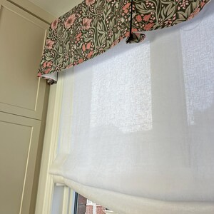 May include: White sheer roman shade with a floral valance in shades of pink and green. The valance has a pleated design.