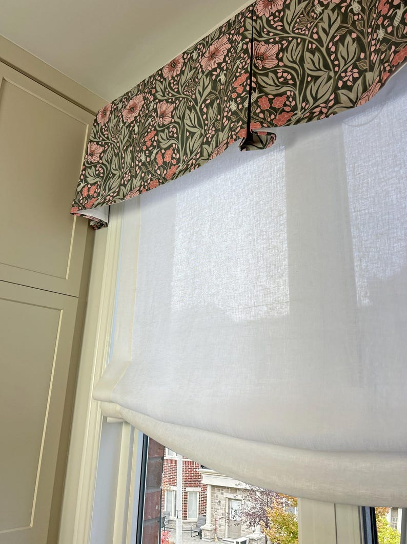 May include: White sheer roman shade with a floral valance in shades of pink and green. The valance has a pleated design.