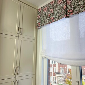 May include: A white roman shade with a floral valance in shades of pink and green hangs in a window. The window is framed in white and looks out onto a brick building with trees in the background.