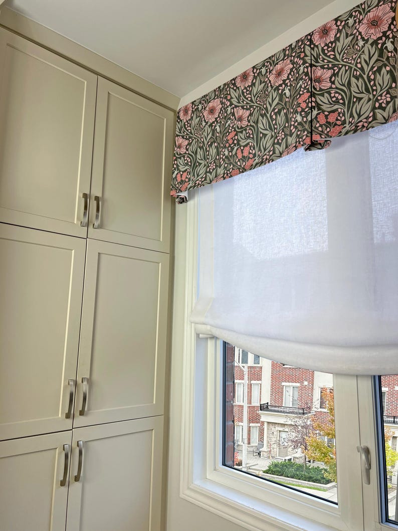 May include: A white roman shade with a floral valance in shades of pink and green hangs in a window. The window is framed in white and looks out onto a brick building with trees in the background.