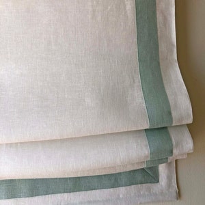 May include: White linen Roman shade with a green trim. The shade is folded up, showing the layers of fabric.