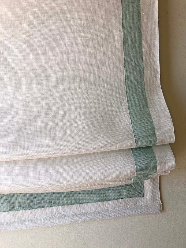 May include: White linen Roman shade with a green trim. The shade is folded up, showing the layers of fabric.