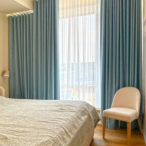 May include: Floor-to-ceiling blue curtains with a textured fabric finish, paired with sheer white vertical pleated drapes. The curtains hang from a track system covering a large window, creating a layered window treatment with soft folds.