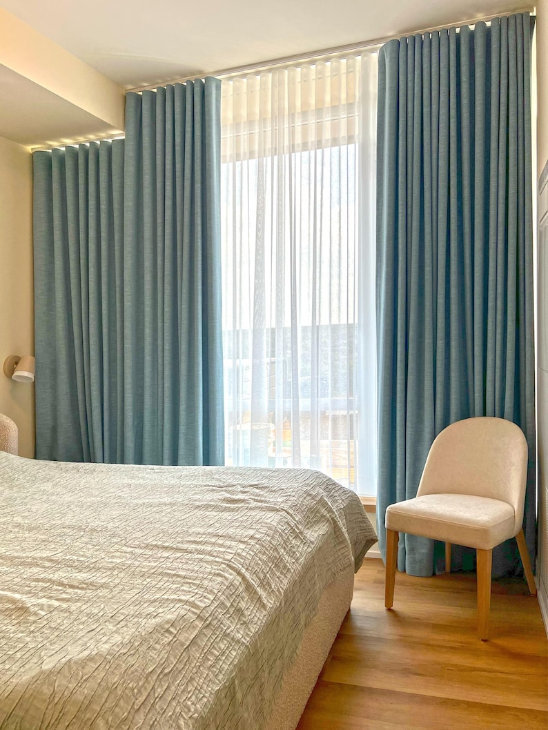 May include: Floor-to-ceiling blue curtains with a textured fabric finish, paired with sheer white vertical pleated drapes. The curtains hang from a track system covering a large window, creating a layered window treatment with soft folds.